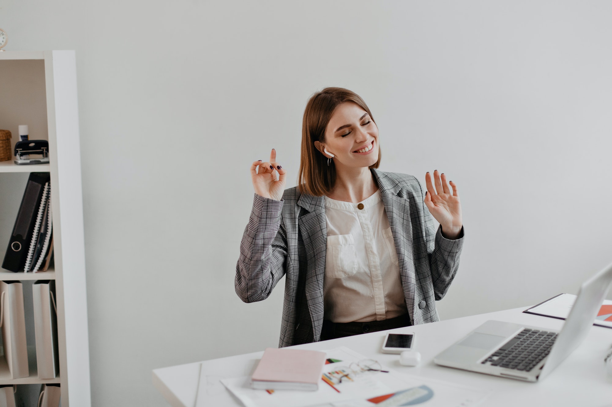 Business woman in gray jacket enjoying music while sitting at workplace in white office