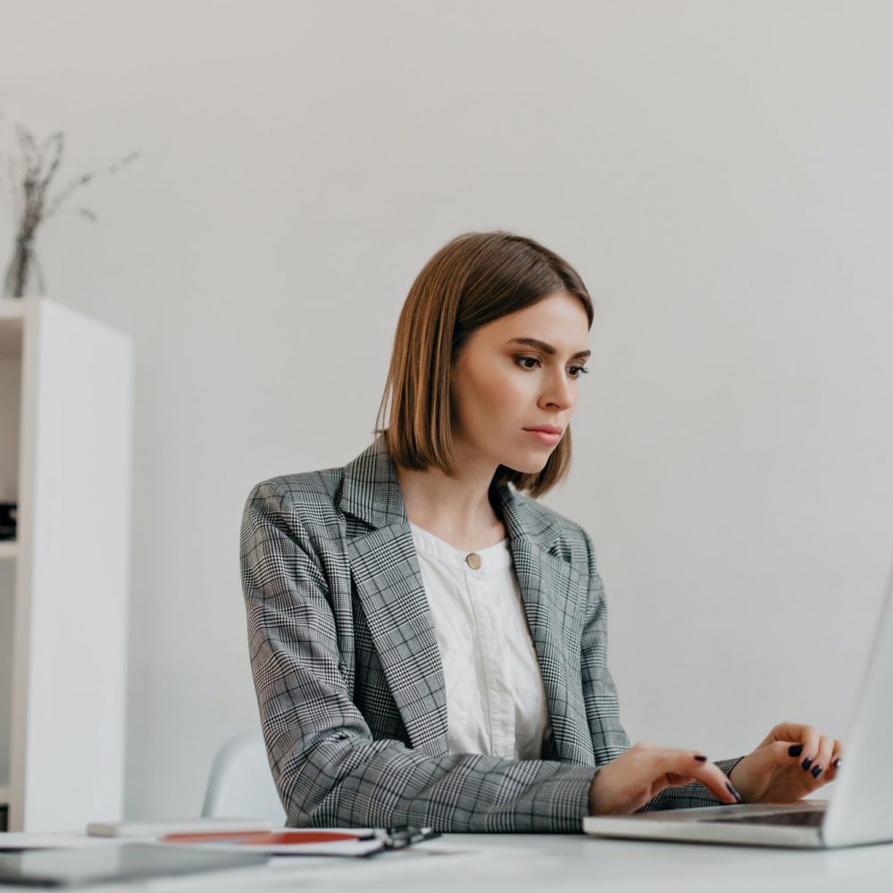 Attractive blonde woman typing letter in MacBook at her workplace. Photo of lady in stylish jacket