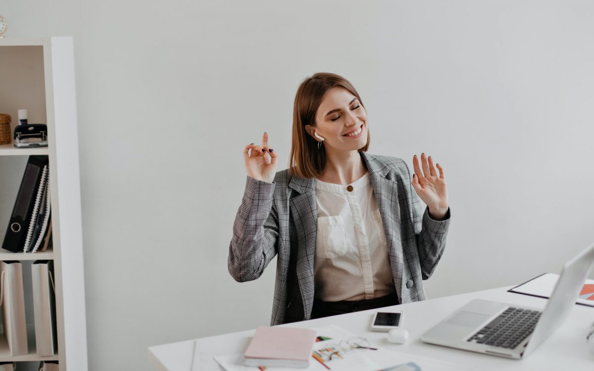 Business woman in gray jacket enjoying music while sitting at workplace in white office